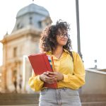 Smiling female student with curly hair and glasses holding books on campus steps.