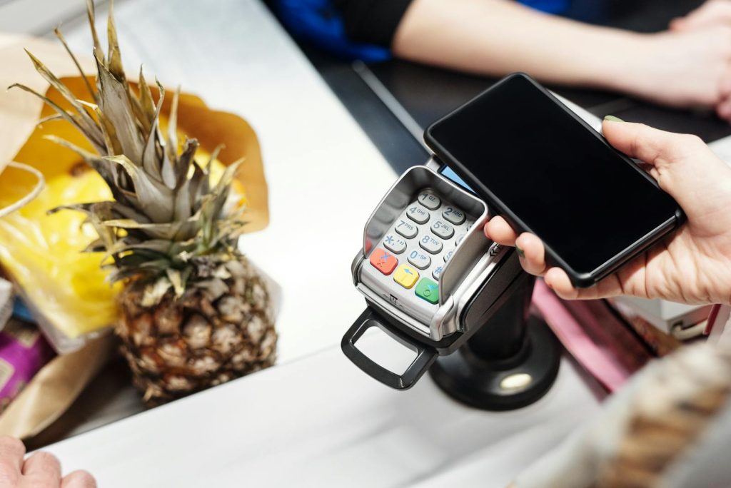 Close-up of a customer using a smartphone for contactless payment at a retail checkout with a pineapple on the counter.