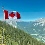 Canadian flag waving atop a scenic mountain view with blue skies and lush greenery.