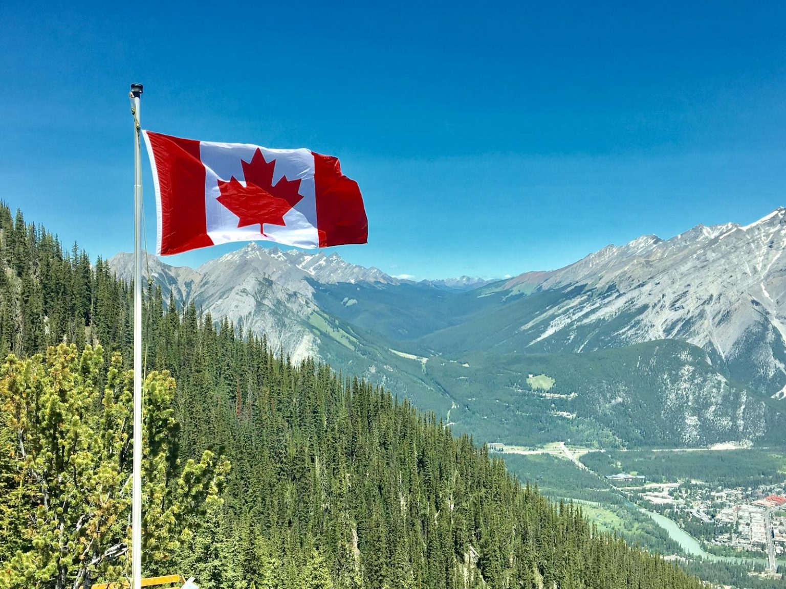 Canadian flag waving atop a scenic mountain view with blue skies and lush greenery.